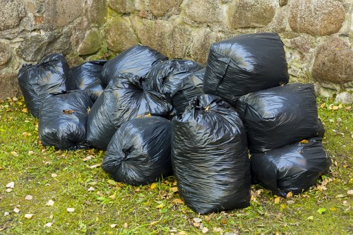 A collection of ten black plastic rubbish bags, tightly sealed with knot closures, are grouped together on a patch of grass in an outdoor setting. The bags are placed close to a rough stone wall, with some bags leaning against each other and others standing upright. The grass beneath is slightly uneven and interspersed with small scattered leaves. The bags are made of glossy black plastic, reflecting natural light, and appear to contain waste materials typical of rubbish removal services offered by Office Clearance Stockwell. The stone wall behind them exhibits a textured, weathered surface with various shades of brown and gray, indicating an outdoor environment possibly adjacent to a property in Stockwell or the surrounding London area. The scene conveys a straightforward view of waste ready for collection or disposal, aligned with the company's workplace clearance and rubbish removal offerings in the local area.