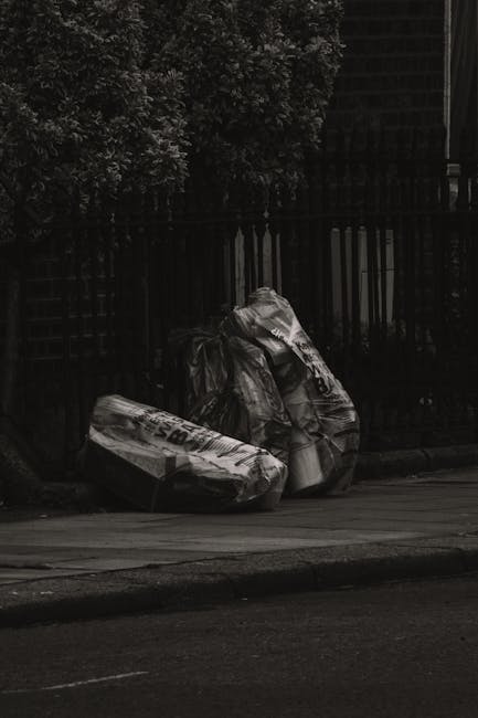 Two large bags of mixed rubbish, one plastic and one paper, are placed on the pavement near a black metal fence. The plastic bag appears filled with waste materials, with some items protruding from th