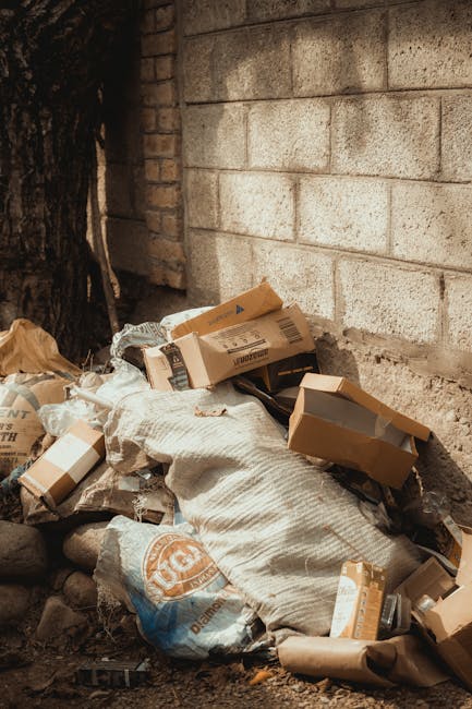 Two large bags of mixed rubbish, one plastic and one paper, are placed on the pavement near a black metal fence. The plastic bag appears filled with waste materials, with some items protruding from th