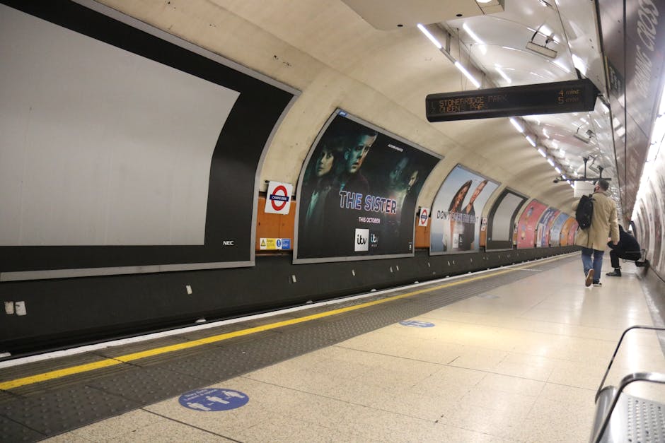 Inside a London underground station, there is a curved tunnel wall with large advertisement posters positioned along the side. The posters include a mix of images and text, such as one promoting a TV