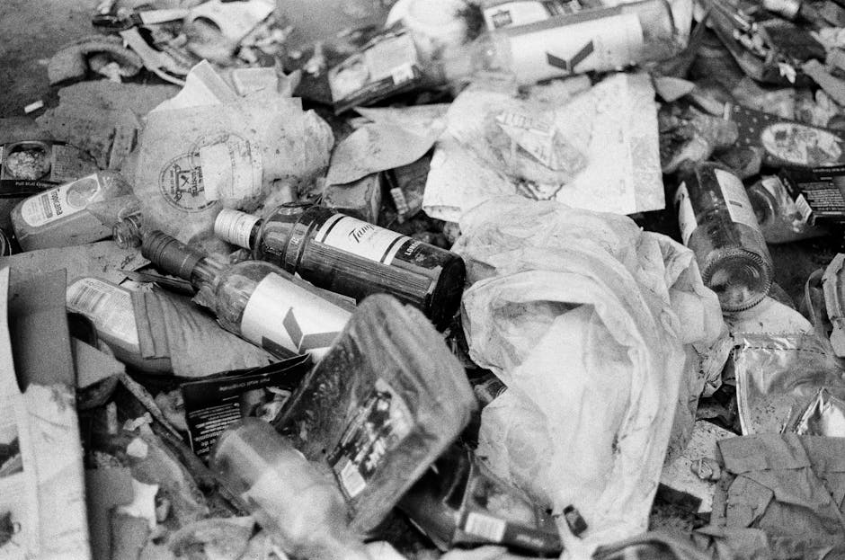 A collection of discarded household waste, including cardboard boxes, plastic containers, a small wooden cabinet, and miscellaneous debris, is scattered among dry, leafless branches and twigs on the g